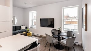 A modern living room with a sofa, a dining table, and chairs. There is a flat-screen TV mounted on the wall, and large windows providing natural light.