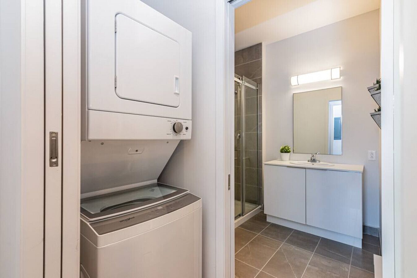 Ein moderner Waschraum mit einer Waschmaschine und einem Trockner, einer Duschkabine aus Glas sowie ein Badezimmer mit Waschbecken und Spiegel.