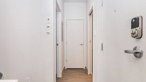 A narrow hallway with white walls, a wooden floor, and a door at the end. There are two wall-mounted devices on the right side, one appears to be a door lock and the other a security camera.