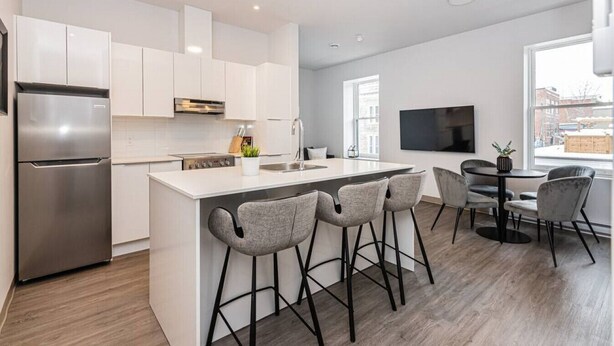 A modern kitchen with white cabinetry, a stainless steel refrigerator, a central island with bar stools, and a dining area with a round table and chairs.
