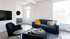 A modern living room with a blue sofa, black armchairs, a coffee table, a flat-screen TV, and a large window. - Stunning 1 Bedroom in Mile End by Denstays (Montreal)