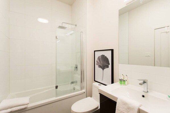 A modern bathroom with a glass-enclosed shower, a toilet, a sink with toiletries, and a framed artwork on the wall. - Stunning 1 Bedroom in Mile End by Denstays (Montreal)