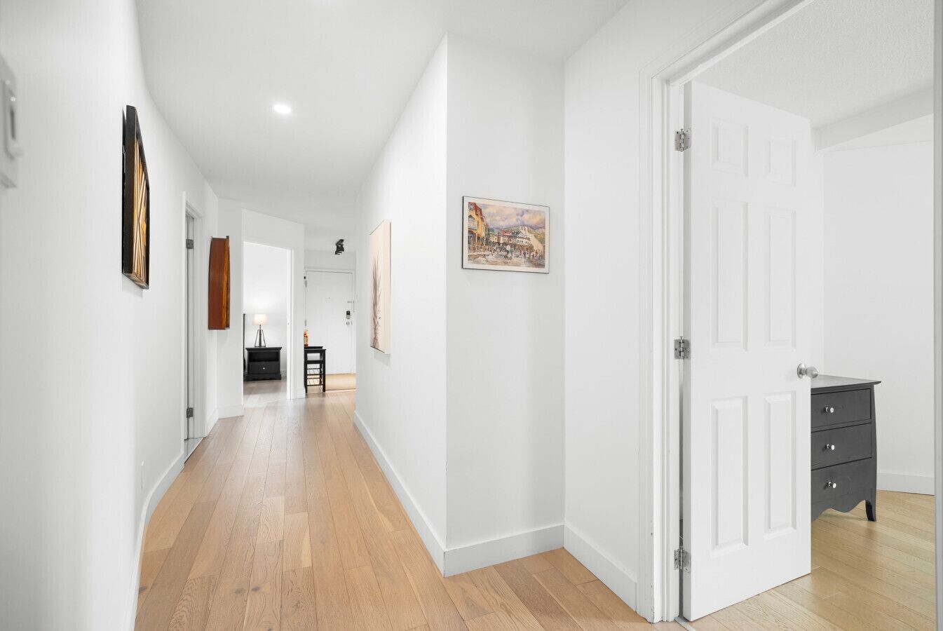 A hallway with wooden flooring, white walls, and a door with a dark dresser.