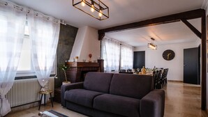 Un salon avec un canapé foncé, une table basse en verre et une cheminée. On y trouve également un coin repas avec une table dressée pour six personnes et une horloge au mur.