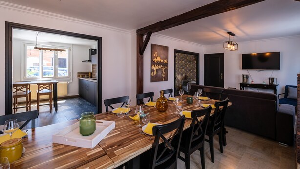 Un coin repas avec une table en bois dressée pour six personnes, une cuisine en arrière-plan et un téléviseur fixé au mur.