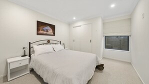 A bedroom with a bed, nightstand, and a window with a view of a wooden deck. - Kapiti Views - Waikanae Beach Holiday Home (Waikanae Beach)
