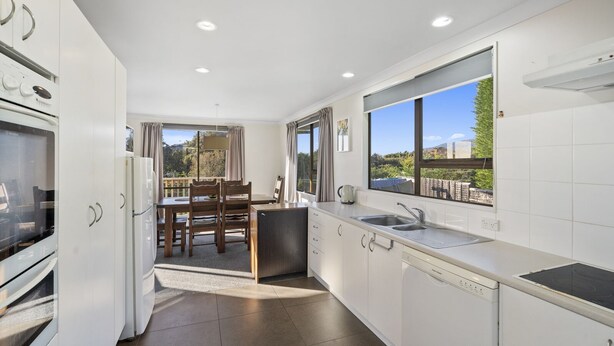 A modern kitchen with white cabinetry, a built-in oven, a sink, and a dining area with a table and chairs. - Kapiti Views - Waikanae Beach Holiday Home (Waikanae Beach)