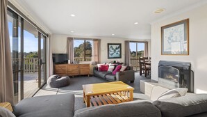 A spacious living room with a grey sofa, a wooden coffee table, and a fireplace. There is a television on a wooden cabinet and a balcony visible through large windows. - Kapiti Views - Waikanae Beach Holiday Home (Waikanae Beach)