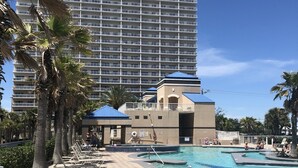 Zona de piscina hotelera con sillones, palmeras y un edificio grande al fondo.