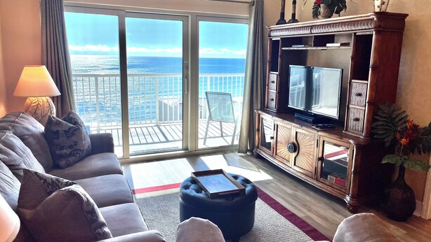 Un salón con sofá, reposapiés y televisor sobre un mueble de madera. Se ve un balcón con vistas al océano a través de puertas de cristal.