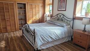 Een slaapkamer met een bed, houten vloer, plafondventilator, een houten kledingkast en een raam met uitzicht.