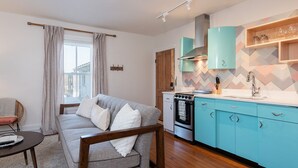 A modern living room with a grey sofa, wooden coffee table, and a window with curtains. Adjacent is a kitchen with teal cabinets, a stove, and a sink. - Retro Apartment | Furnished Monthly Rental in Belmont (Charlottesville)