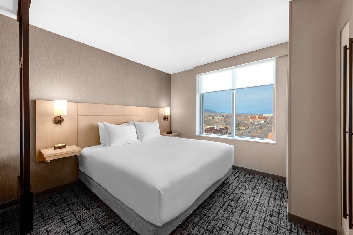 Habitación de hotel con una cama grande, dos lámparas de noche, una mesita de noche y una ventana con vistas a la ciudad.