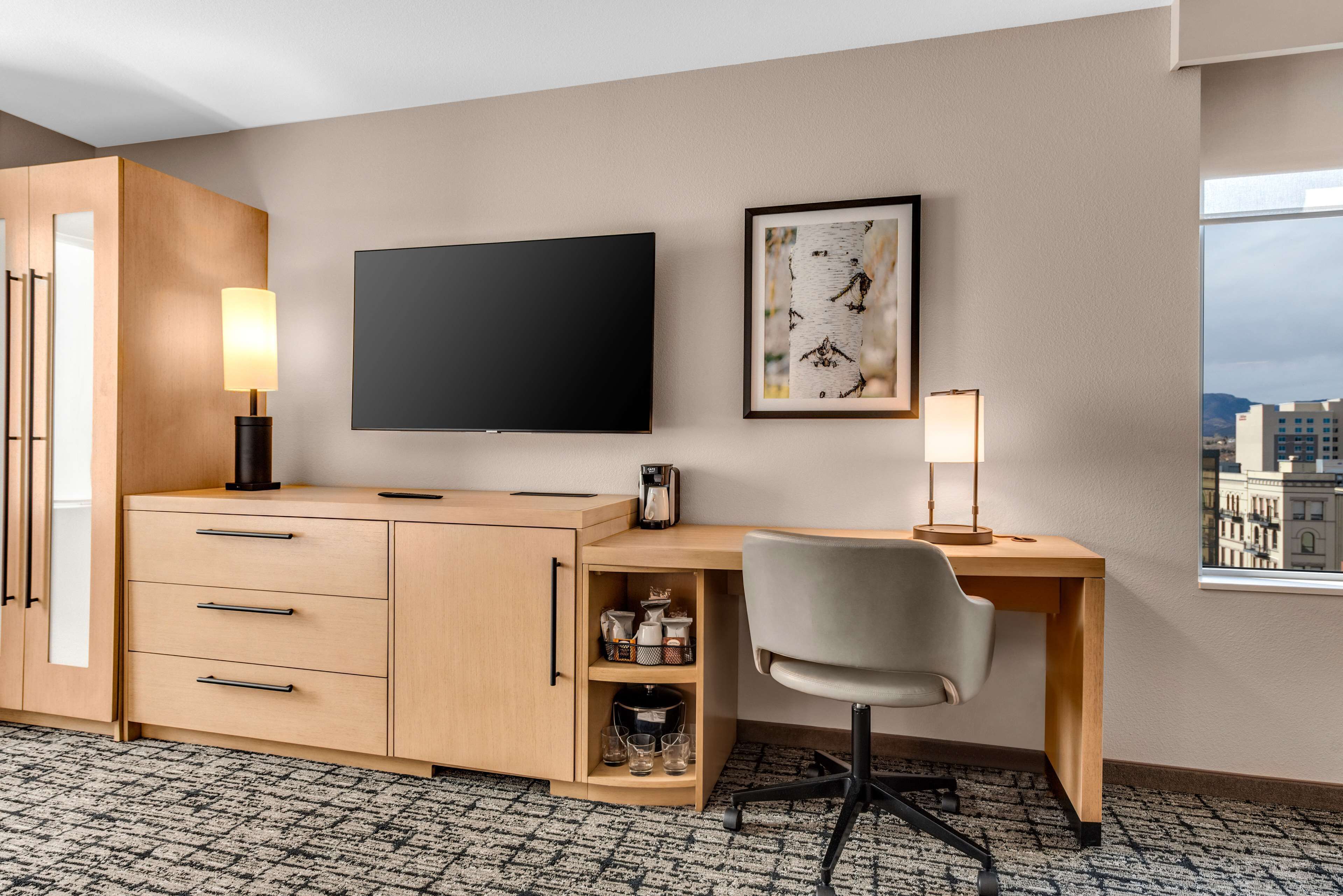 A hotel room with a wooden desk, a flat-screen TV, a framed picture on the wall, and a window with a city view.