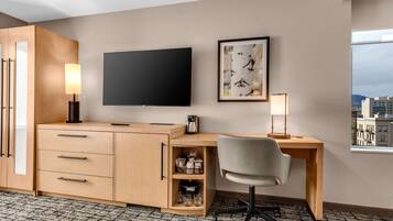 A hotel room with a wooden desk, a flat-screen TV, a framed picture on the wall, and a window with a city view.