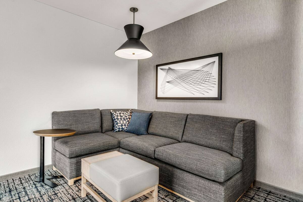 Una sala de estar moderna con un sofá seccional gris, una mesita redonda pequeña, un reposapiés de color claro, una lámpara colgante negra y una obra de arte abstracta enmarcada en la pared.