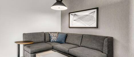 A modern living room with a gray sectional sofa, a small round table, a light-colored ottoman, a black pendant light, and a framed abstract artwork on the wall.