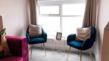 Ein Zimmer mit zwei blauen Sesseln, einem kleinen Beistelltisch mit einem 'WELCOME'-Schild, einem Fenster mit Vorhängen und einem roten Sofa mit einem bunten Kissen.