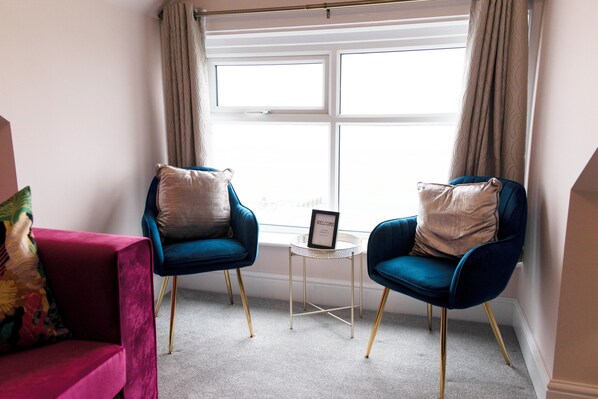 Une pièce avec deux fauteuils bleus, une petite table d’appoint sur laquelle est posé un panneau indiquant « BIENVENUE », une fenêtre avec des rideaux et un canapé rouge agrémenté d’un coussin coloré.