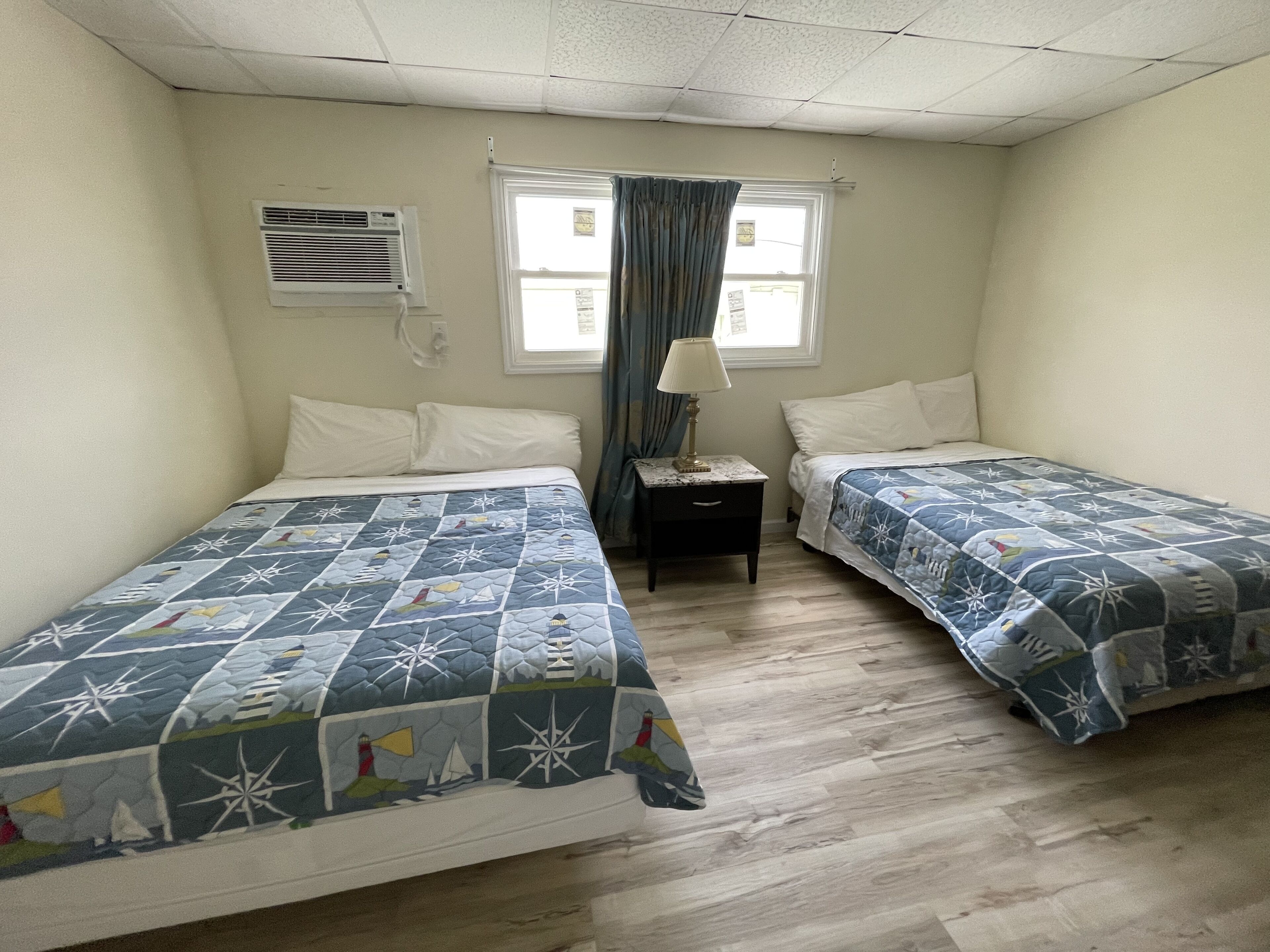Une chambre avec deux lits, chacun recouvert d’une courtepointe matelassée, une table de chevet avec une lampe, une fenêtre avec des rideaux et un climatiseur fixé au mur.