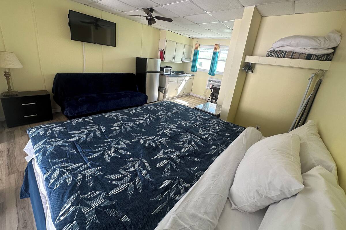 Habitación de hotel compacta con cama, escritorio, silla, televisor, ventilador de techo y kitchenette.