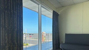 A room with a large sliding glass door leading to a balcony, a dark-colored sofa, and a view of the street and ocean. - Searay Motel (Wildwood)