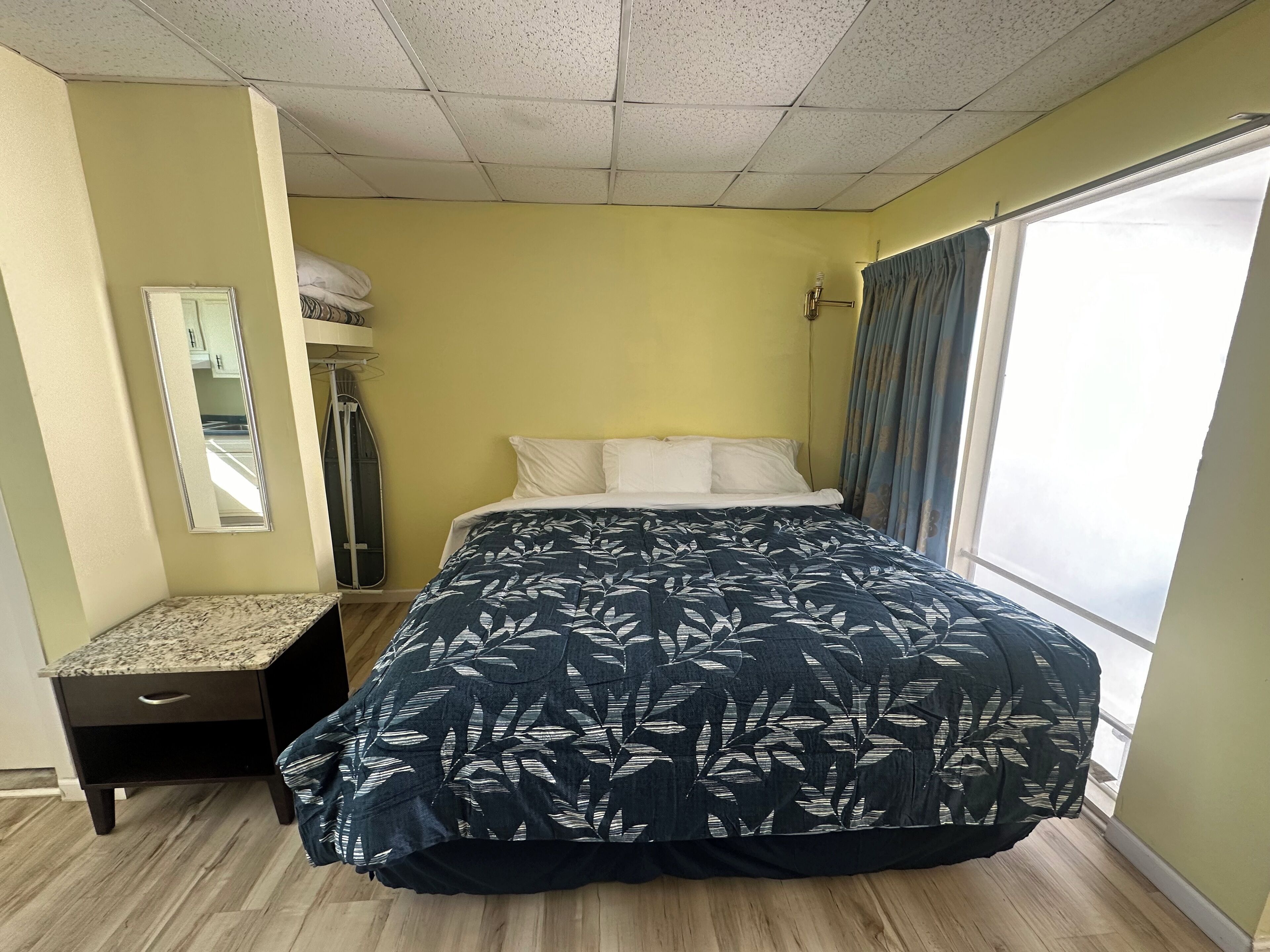 A bedroom with a bed, a nightstand, a mirror, and a window with curtains.