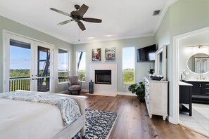 A bedroom with a bed, a ceiling fan, a fireplace, a TV, and a large window.