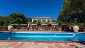 Une piscine avec des chaises longues et des parasols, une terrasse en pierre et un bâtiment en arrière-plan.