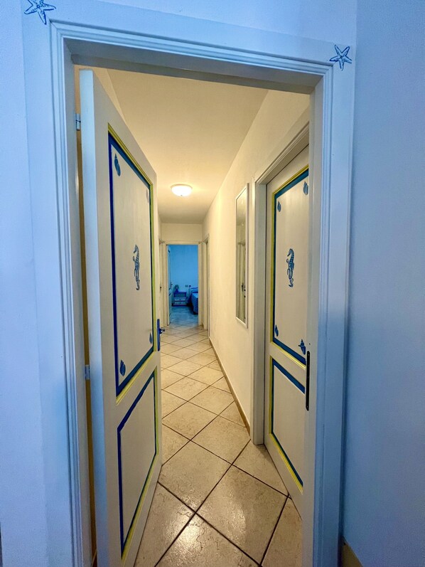 Um corredor com duas portas, cada uma adornada com um emblema decorativo. O chão é revestido com azulejos e as paredes pintadas em tom claro.