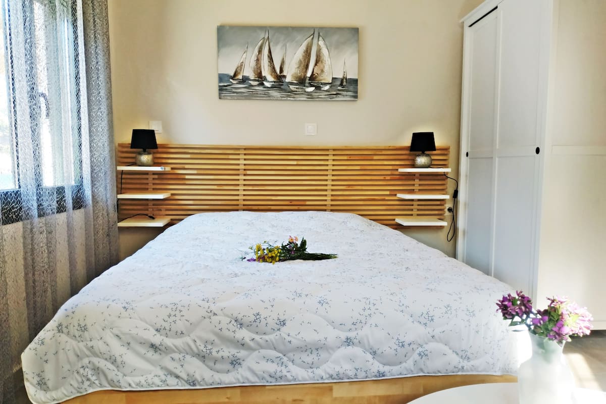 Een slaapkamer met een bed, een nachtkastje, een lamp, een schilderij van zeilboten en een deur.