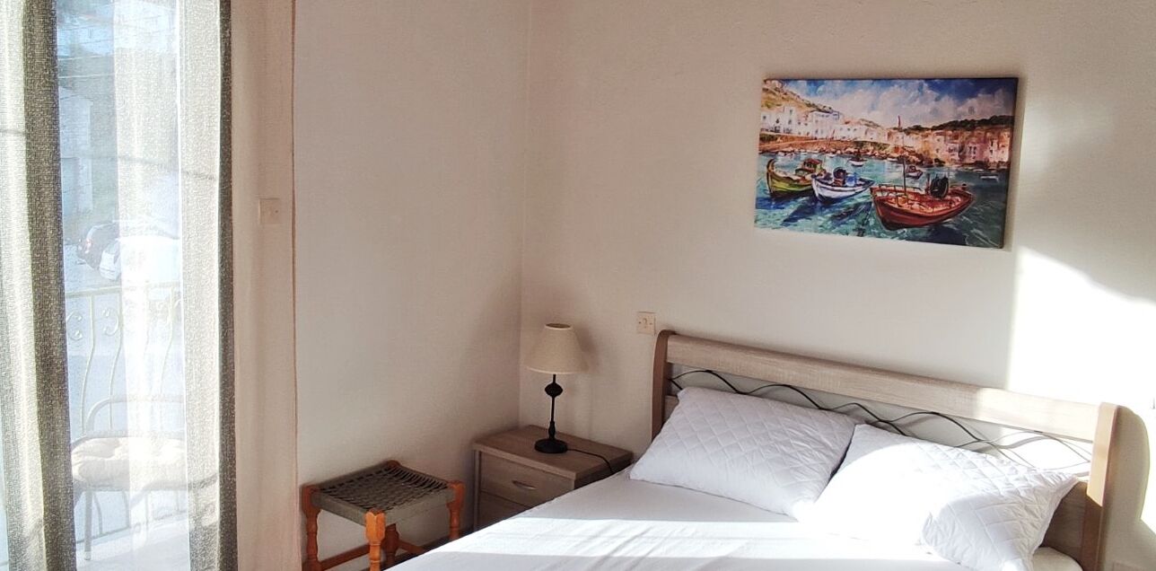 A hotel room with a bed, a nightstand with a lamp, a small table, and a window with curtains.