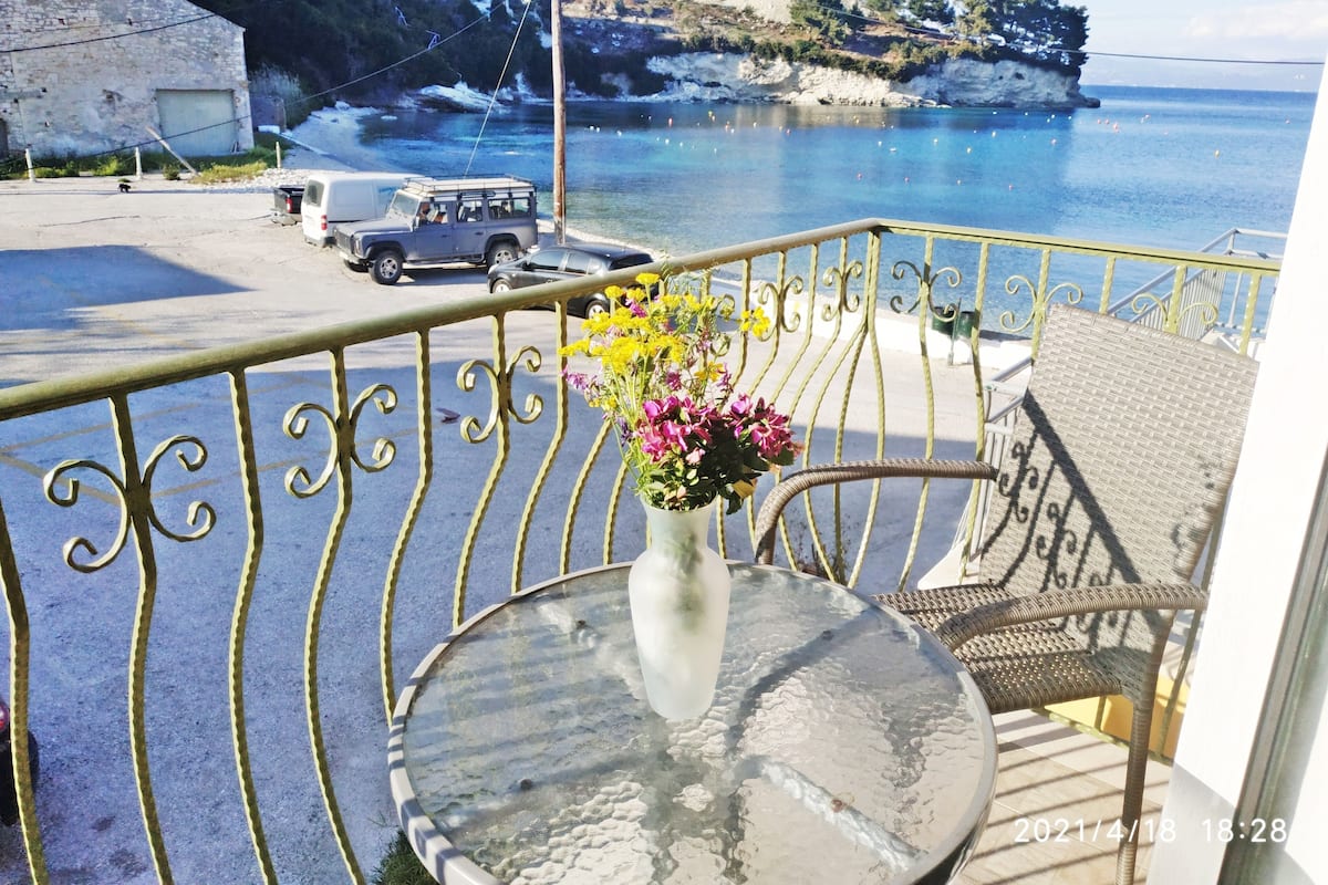 Een balkon met een metalen hekwerk, een tafel met een bloevas, en uitzicht op zee en een geparkeerde auto.