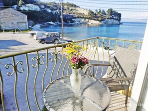 Un balcón con barandilla de metal, una mesa con un jarrón de flores y vista al mar, con un coche estacionado.