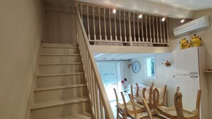 A staircase with a wooden handrail leading to a second-floor balcony. A dining area with a table and chairs is visible below. - Agréable Studio Dans Jardin Paysager Avec Piscine et Grande Cuisine D'été (Bagnols-en-Forêt)