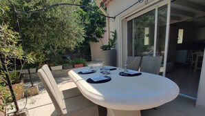 A round table with four chairs, a glass door leading to an indoor area, and potted plants around. - Agréable Studio Dans Jardin Paysager Avec Piscine et Grande Cuisine D'été (Bagnols-en-Forêt)