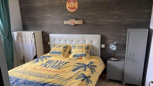 A bedroom with a bed featuring a 'Wave Patrol California' themed bedspread, a wooden wall with a 'Gasoline Route 66 Top Quality' sign, a grey wardrobe, and a nightstand with a lamp. - Gîte "prima Luce" (Preures)