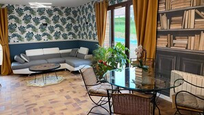 A living room with a sectional sofa, a glass coffee table, wicker chairs, a bookshelf, and a patterned wallpaper. - Gîte "prima Luce" (Preures)