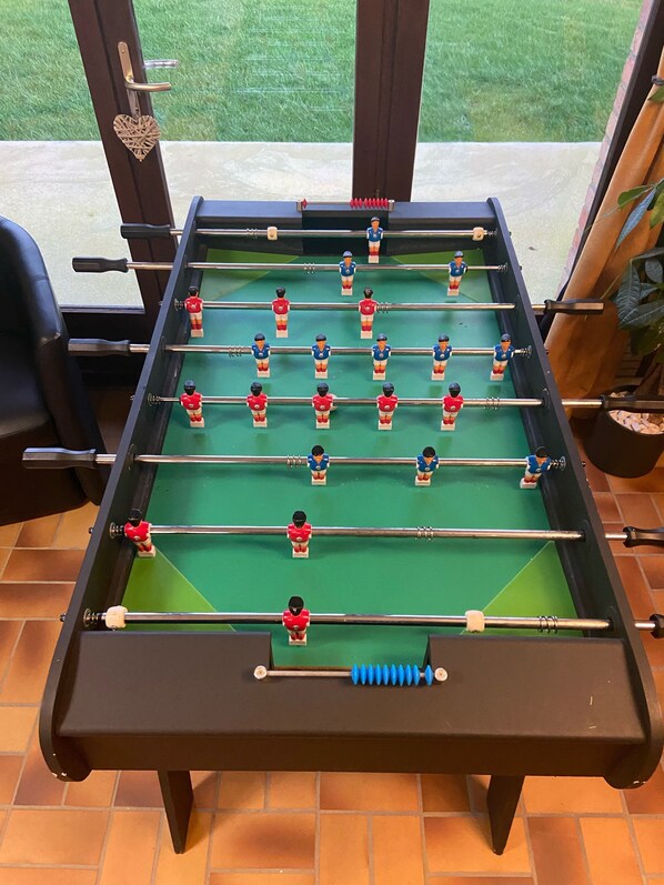 A foosball table with a heart-shaped decoration on the door, a view of a grassy area through the window, and a tiled floor.