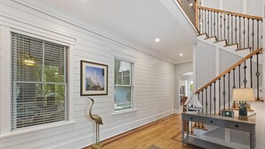Un couloir aux murs recouverts de panneaux blancs, doté d’un plancher en bois, d’un escalier avec des balustrades noires, et d’un bureau muni de tiroirs et d’une lampe.