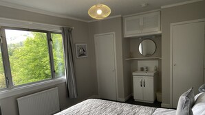 Um quarto com cama, janela com cortinas, radiador, guarda-roupa, espelho e uma pequena prateleira com artigos de higiene pessoal.