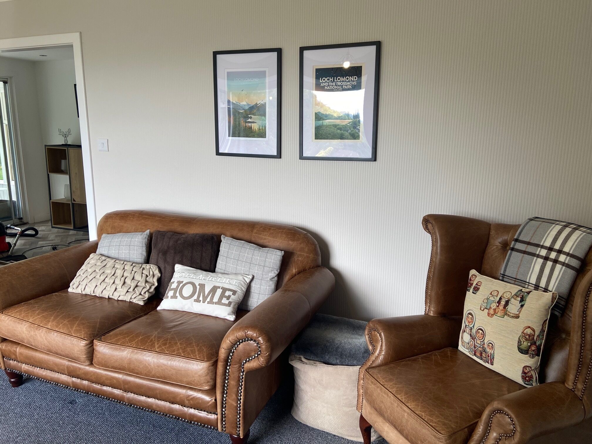 Un sofá de cuero marrón con un cojín que dice 'NO PLACE LIKE HOME' y un sillón a juego con un almohadón de cuadros. Dos cuadros enmarcados de Loch Lomond y el Parque Nacional de las Tierras Altas Escocesas están colgados en la pared.
