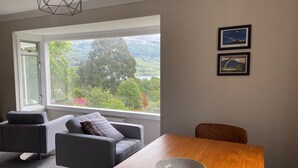 Quarto moderno com uma janela grande que oferece uma vista panorâmica de árvores e colinas. Há uma mesa de jantar de madeira com uma tigela sobre ela, duas cadeiras cinzas e um pequeno sofá com uma almofada.