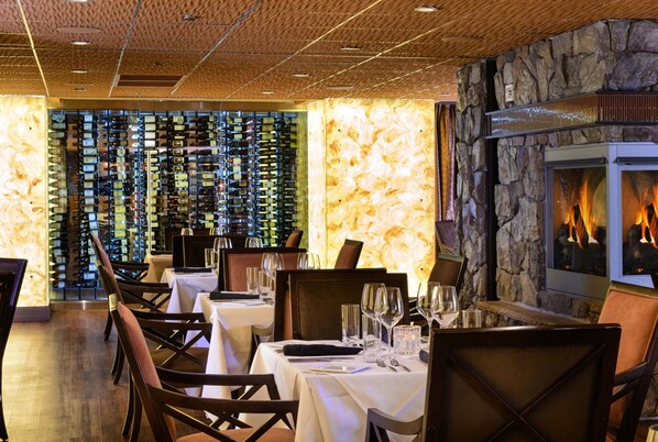 Un restaurant avec un mur en pierre et une cheminée, des tables dressées avec des verres à vin et des couverts, et un mur entier recouvert de bouteilles de vin.