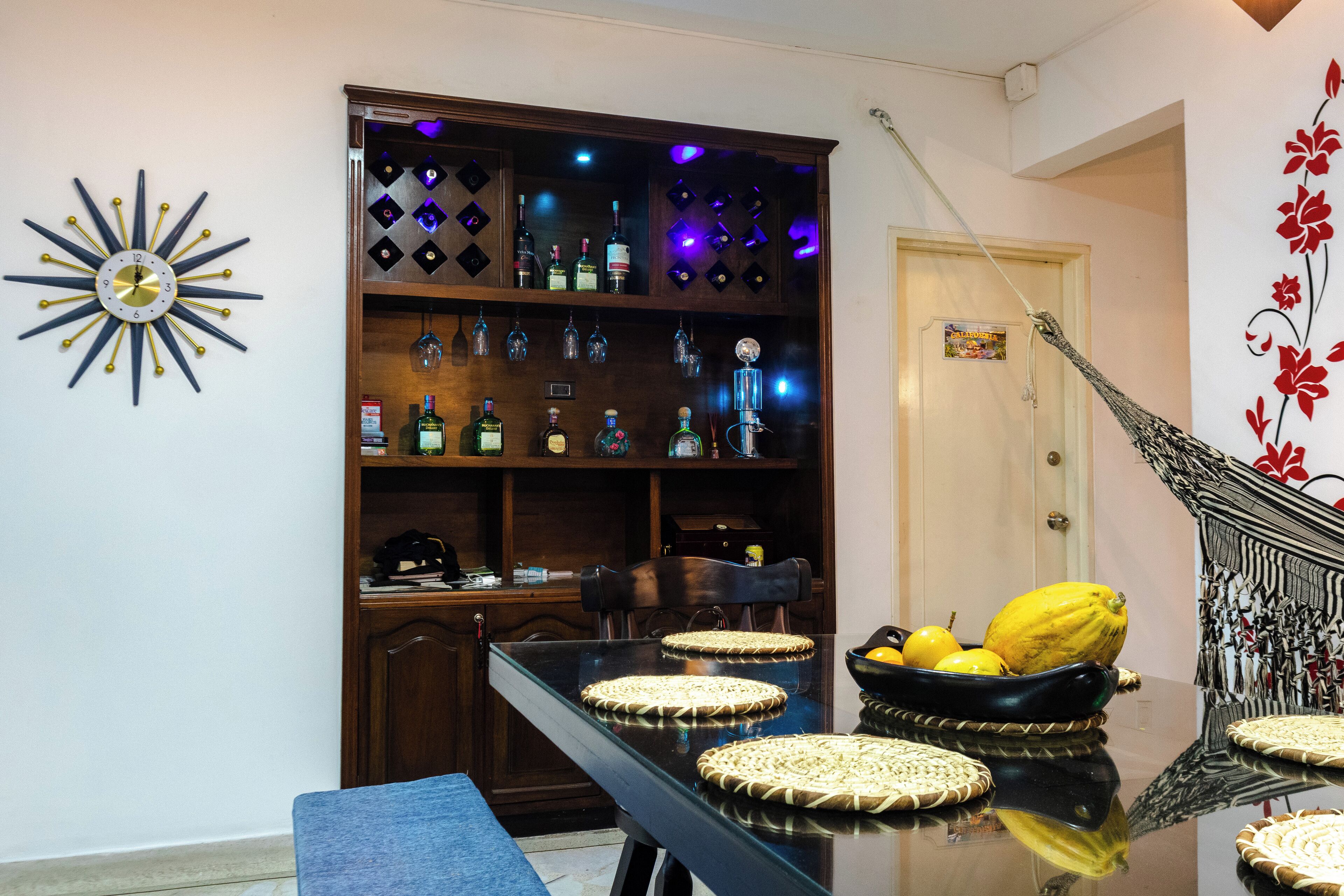 A dining area with a glass table, woven placemats, and a decorative wall clock.