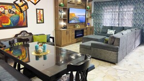 A modern living room with a dining area, featuring a wooden entertainment unit, a flat-screen TV, and a large sofa.