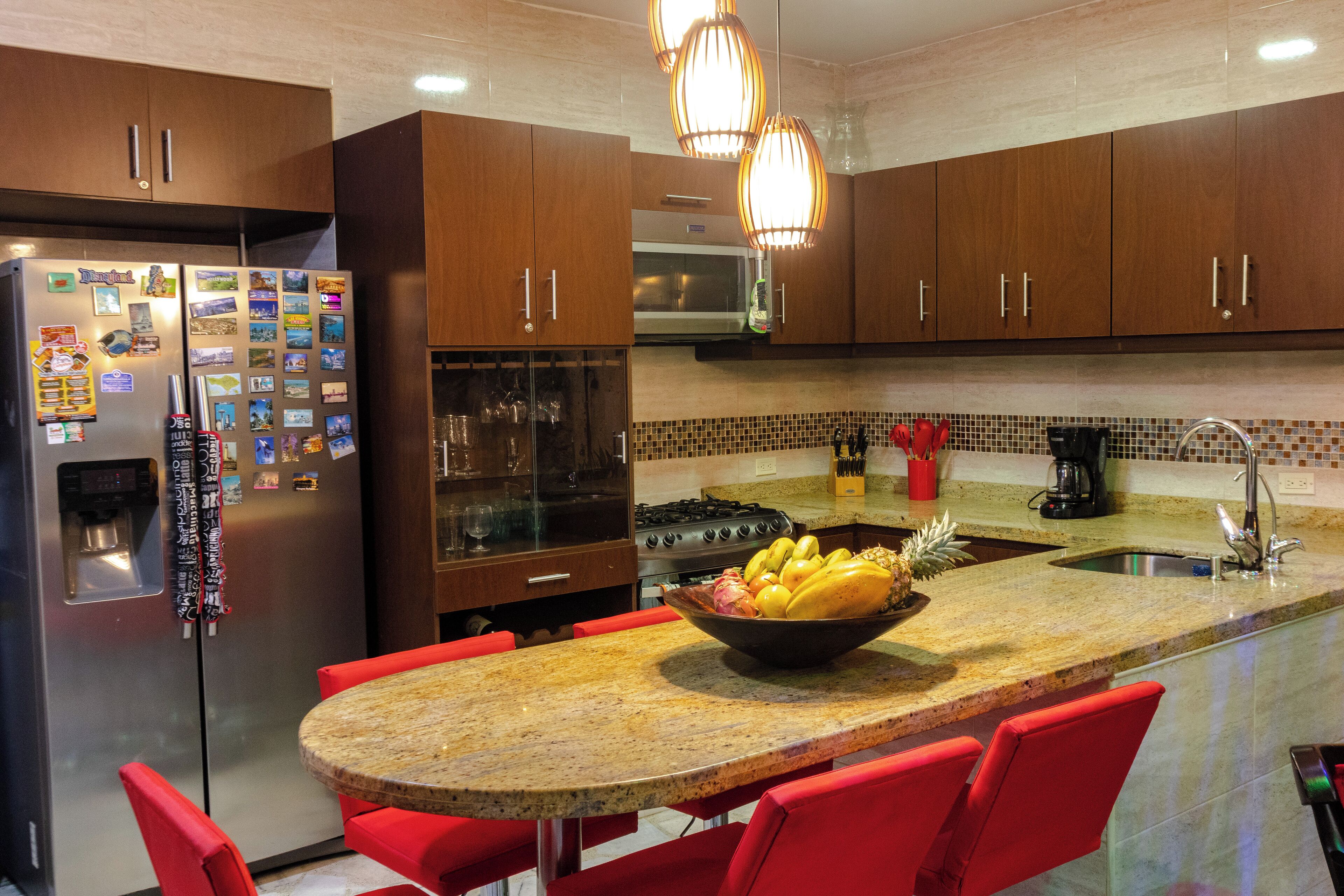 A modern kitchen with wooden cabinets, a stainless steel refrigerator, a granite countertop, and red bar stools.