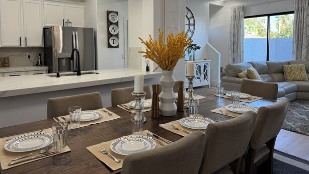 A modern kitchen with white cabinets, a stainless steel refrigerator, and a dining area set for six with a large table, chairs, and a chandelier.