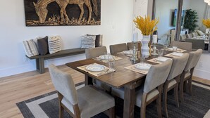 A dining area with a wooden table set for six, surrounded by grey upholstered chairs. A large wooden horse sculpture hangs above the table, and a mirror reflects the room.