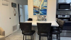 A modern kitchen with a central island, bar stools, a painting of a sailboat, and a sign reading "Our Kitchen". - Wave Action!Condo on the Beach w/Umbrella Service next to famous Wanna Wanna Bar (South Padre Island)